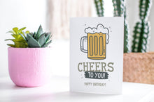 Load image into Gallery viewer, A greeting card featured standing up on a white tabletop with a pink plant pot in the background and some succulents in the pot. There’s a woven basket in the background with a cactus inside. The card features the words “Cheers to You! Happy Birthday!” with an illustrated beer mug.
