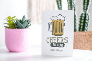A greeting card featured standing up on a white tabletop with a pink plant pot in the background and some succulents in the pot. There’s a woven basket in the background with a cactus inside. The card features the words “Cheers to You! Happy Birthday!” with an illustrated beer mug.