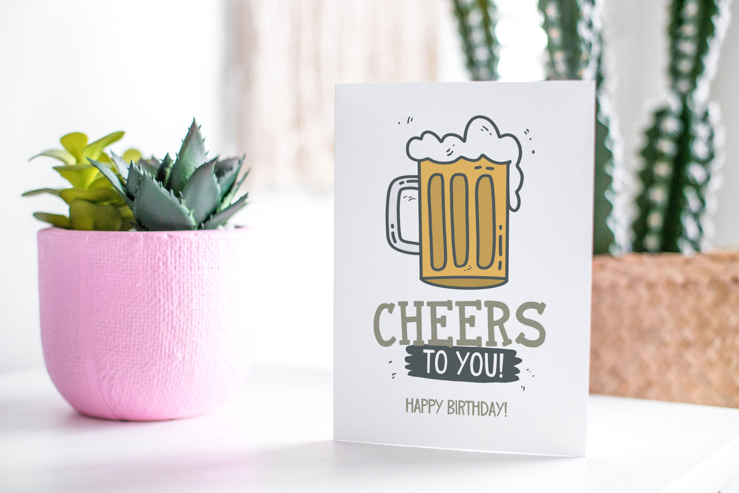 A greeting card featured standing up on a white tabletop with a pink plant pot in the background and some succulents in the pot. There’s a woven basket in the background with a cactus inside. The card features the words “Cheers to you, happy birthday” with an illustrated beer mug.