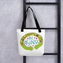 Load image into Gallery viewer, A white tote bag with black handles handing on a black ladder. The bag features a design with the words &#39;be kind&#39; with illustrated hugging hands around the words. 