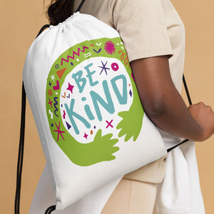 A woman holding a white drawstring bag. The drawstring bag has the words "Be Kind" with green illustrated hands hugging the design with sketchy illustrations. 