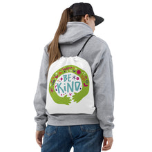 Load image into Gallery viewer, A woman holding a white drawstring bag on her back. She&#39;s wearing a grey hoodie and a black hat. The drawstring bag has the words &quot;Be Kind&quot; with green illustrated hands hugging the design with sketchy illustrations. 