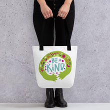 Load image into Gallery viewer, A person holding a white tote bag with black handles. The bag features a design with the words &#39;be kind&#39; with illustrated hugging hands around the words. 