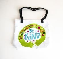 Load image into Gallery viewer, A white tote bag with black handles on a white tabletop. The bag features a design with the words &#39;be kind&#39; with illustrated hugging hands around the words. 