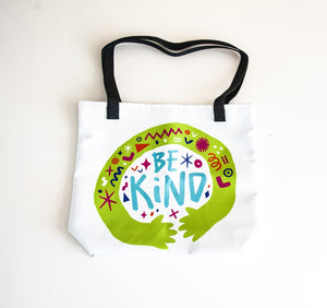 A white tote bag with black handles on a white tabletop. The bag features a design with the words 'be kind' with illustrated hugging hands around the words. 