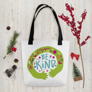 A white tote bag with black handles on a wood tabletop with Christmas items around it. The bag features a design with the words 'be kind' with illustrated hugging hands around the words. 