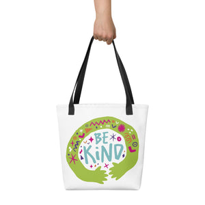 Someone holding a white tote bag with black handle. The bag features a design with the words 'be kind' with illustrated hugging hands around the words. 