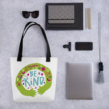 Load image into Gallery viewer, A white tote bag with black handles on a grey tabletop with a laptop and other office supplies. The bag features a design with the words &#39;be kind&#39; with illustrated hugging hands around the words. 