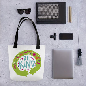 A white tote bag with black handles on a grey tabletop with a laptop and other office supplies. The bag features a design with the words 'be kind' with illustrated hugging hands around the words. 