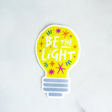 Load image into Gallery viewer, A die cut vinyl sticker with the words &quot;be the light&quot; inside an illustrated lightbulb.