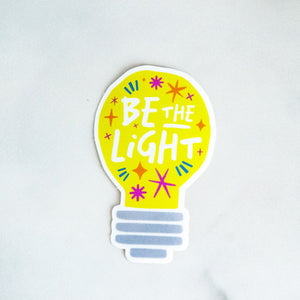 A die cut vinyl sticker with the words "be the light" inside an illustrated lightbulb.