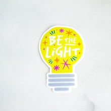 Load image into Gallery viewer, A die cut vinyl sticker with the words &quot;be the light&quot; inside an illustrated lightbulb.