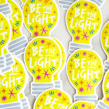 Load image into Gallery viewer, Die cut vinyl stickers with the words &quot;be the light&quot; inside an illustrated lightbulb.