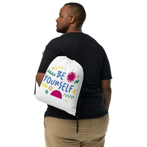 A man holding a white drawstring bag on his shoulder. The drawstring bag has the words "Be Yourself" in blue with green illustrated sketchy pieces around the words. 