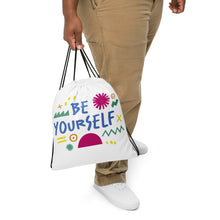 Load image into Gallery viewer, A person holding a white drawstring bag with their hands. The drawstring bag has the words &quot;Be Yourself&quot; in blue with green illustrated sketchy pieces around the words. 