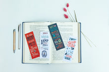 Load image into Gallery viewer, A set of four bookmarks laying across an open Bible. There are two pens off to the lefthand side and dried flowers across the upper righthand corner. 