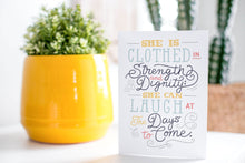 Load image into Gallery viewer, A greeting card is on a table top with a yellow plant pot and a green plant inside. The card features the words &quot;She is clothed in strength and dignity; she can laugh at the days to come.&quot;