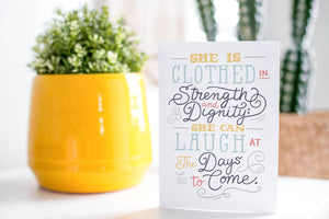 A greeting card is on a table top with a yellow plant pot and a green plant inside. The card features the words "She is clothed in strength and dignity; she can laugh at the days to come."