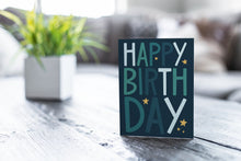 Load image into Gallery viewer, A greeting card featured on a black, wood coffee table. There’s a white planter in the background with a green plant. There’s also a gray sofa in the background with a white pillow. The card features the words “happy birthday” with stars surrounding the letters.