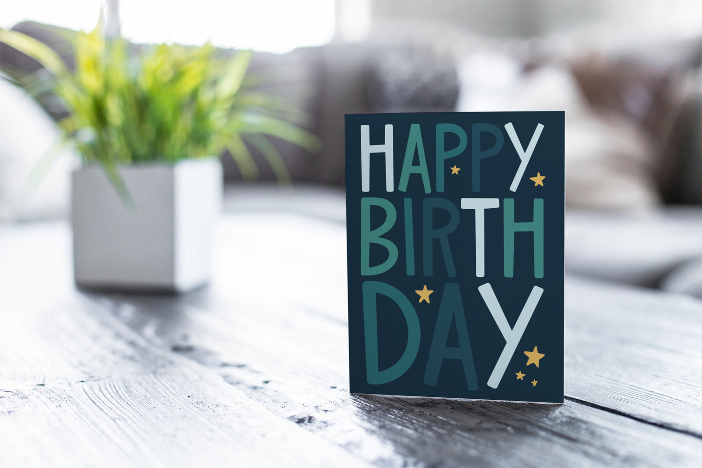 A greeting card featured on a black, wood coffee table. There’s a white planter in the background with a green plant. There’s also a gray sofa in the background with a white pillow. The card features the words “happy birthday” with stars surrounding the letters.