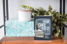 Load image into Gallery viewer, A greeting card is on a table top with a present in blue wrapping paper in the background. On top of the present is a candle and some greenery from a plant too. The card features the words “Have Your Cake and Eat it All Happy Birthday” with an illustrated piece of cake with a candle in it.