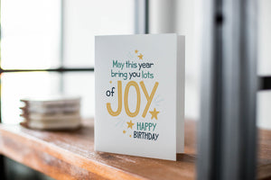 A greeting card featured standing up on a white tabletop with a framed photo of a succulent in the background and a stack of wooden coasters. There’s a woven basket in the background with a cactus inside. The card features the words “May This Year Bring You Lots of Joy, Happy Birthday.”