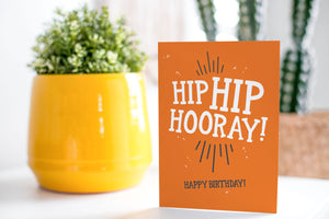 A greeting card is on a table top with a yellow plant pot and a green plant inside. The card features the words “Hip Hip Hooray, Happy Birthday.”