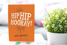 Load image into Gallery viewer, A greeting card is on a table top with a gift in pink wrapping paper. Next to the gift is a white plant pot with a green plant. The card features the words “Hip Hip Hooray, Happy Birthday.”