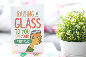 A greeting card is on a table top with a gift in pink wrapping paper. Next to the gift is a white plant pot with a green plant. The card features the words Raising a glass to you on your birthday” with an illustrated hand raising a glass.