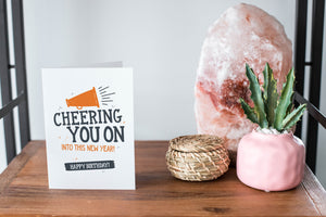 A card on a wood tabletop and on the right side of the card is a woven basket, a pink plant pot with a cactus in it and a pink crystal rock. The card features the words "“Cheering You on into This New Year, Happy Birthday."