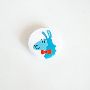 A buttons on a white tabletop. The button features a blue illustrated llama with a red bow tie. 