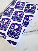 Load image into Gallery viewer, A side view of a spiral notebook is on a tabletop. The white spiral notebook has black spiral binding. The pattern design features the words &quot;I love books&quot; inside an illustrated book with the word love as a heart. 