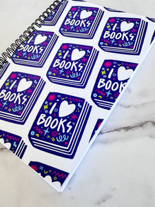 A side view of a spiral notebook is on a tabletop. The white spiral notebook has black spiral binding. The pattern design features the words "I love books" inside an illustrated book with the word love as a heart. 