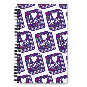 A spiral notebook is on a tabletop. The white spiral notebook has black spiral binding. The pattern design features the words "I love books" inside an illustrated book with the word love as a heart. 