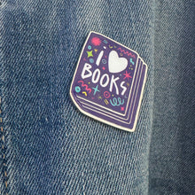 Load image into Gallery viewer, An enamel pin featured on a jean jacket. The pin has the words &#39;I love books&quot; with the word love shown as a heart. The words are featured inside an illustrated book, all in bright fun colors. 