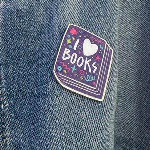 An enamel pin featured on a jean jacket. The pin has the words 'I love books" with the word love shown as a heart. The words are featured inside an illustrated book, all in bright fun colors. 