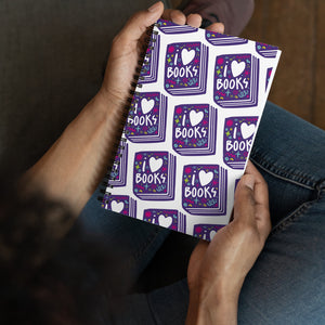 A spiral notebook being held by a person. The white spiral notebook has black spiral binding. The pattern design features the words "I love books" inside an illustrated book with the word love as a heart. 