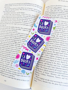 A bookmark placed inside a book. The bookmark features the words "i love books" with love shown shaped as a heart. The words are placed inside an illustrated book. 