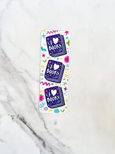 Load image into Gallery viewer, A bookmark on a tabletop. The bookmark features the words &quot;i love books&quot; with love shown shaped as a heart. The words are placed inside an illustrated book. 