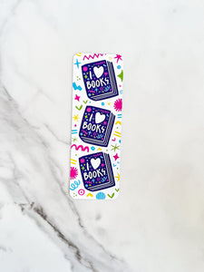 A bookmark on a tabletop. The bookmark features the words "i love books" with love shown shaped as a heart. The words are placed inside an illustrated book. 