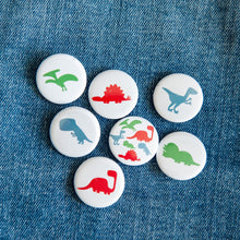 Load image into Gallery viewer, A set of seven buttons on a jean jacket. The buttons feature illustrations of a Pterodactyl, Sauropod, Stegosaurus, T-Rex, Triceratops, Velociraptor, and a button with all on one button. 