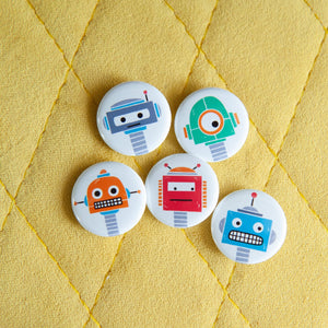 A set of five button pins on a yellow background. The buttons feature five different illustrations of robots.