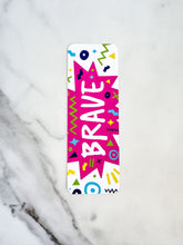 Load image into Gallery viewer, A bookmark with the word &quot;brave&quot; in white within a star-like pink shape with fun, sketchy elemts around. The bookmark is on a tabletop.