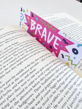 Load image into Gallery viewer, A bookmark with the word &quot;brave&quot; in white within a star-like pink shape with fun, sketchy elemts around. The bookmark is placed inside a book. 