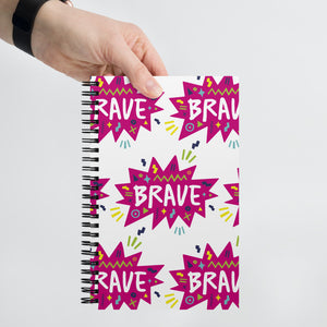 A spiral notebook being held by a person. The white spiral notebook has black spiral binding. The pattern design features the word "brave" inside a hot pink star with sketchy elements around. 