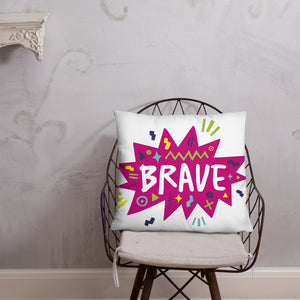 A pillow on a chair against a grey wall. The white pillow features the artwork on a white background with a hot pink star with the word 'brave' inside and sketchy, fun elements around the word. 