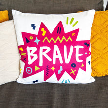 Load image into Gallery viewer, A white pillow on a grey sofa with other pillows. The white pillow features the artwork on a white background with a hot pink star with the word &#39;brave&#39; inside and sketchy, fun elements around the word. 