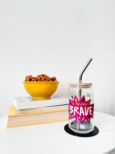 A photo of a glass can tumbler on a coffee table with books and a bowl of pretzels in the background. The glass tumbler has a bamboo lid and silver metal straw. The design on the glass tumbler says the word "brave" inside a hot pink star with illustrated sketchy elements around the word. 