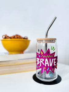 A photo of a glass can tumbler on a coffee table with books and a bowl of pretzels in the background. The glass tumbler has a bamboo lid and silver metal straw. The design on the glass tumbler says the word "brave" inside a hot pink star with illustrated sketchy elements around the word. 