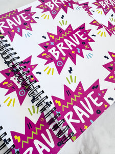 A closeup of three spiral notebooks are on a tabletop. The white spiral notebooks have black spiral binding. The pattern design features the word "brave" inside a hot pink star with sketchy elements around. 